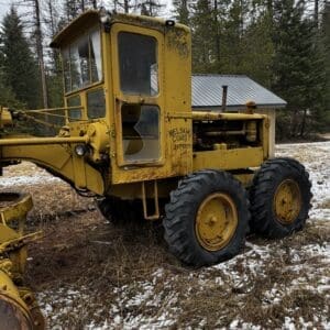 Old yellow construction vehicle with large off-road tires parked in a field with light snow and bare trees in the background