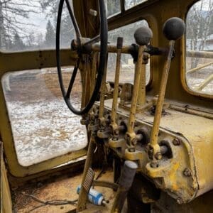 Interior of an old, rusted yellow tractor/cab with a large steering wheel and multiple lever controls in a dusty, snow-dusted environment outside the window.