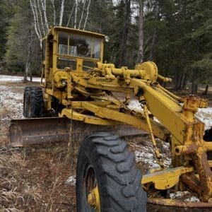 Yellow Caterpillar construction vehicle (grader) in a wooded, lightly snowy area; rust and wear visible on metal frame and blade left in foreground.