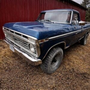 Blue vintage Ford pickup truck with chrome bumper, parked on a dirt surface beside a red wooden fence/barn wall, angled toward the camera.