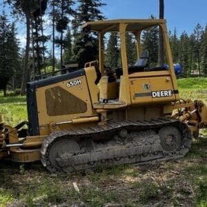 LOT 44 1 - Montana Auctions by Kevin Hill Yellow John Deere 650H LT tracked bulldozer parked on a grassy clearing in a pine forest by a dirt path.