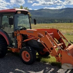 Orange Kubota tractor with front-end loader parked on a gravel path, grassy field and mountains in the background, under a blue sky with clouds.