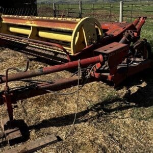 Old red farm implement with a yellow hay baler/rake in a field, rusted and stored near a fence.