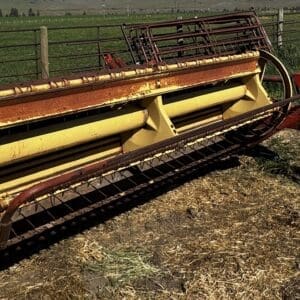 A weathered yellow farm baler sits on dirt near a pasture fence, showing rust and wear in a rural field.