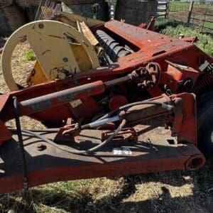 Red, rusted agricultural implement with a large beige reel and exposed gears parked in a grassy yard.