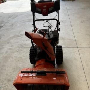 Top-down view of a red snow blower in a garage, with handlebars and controls visible.
