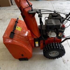 Red snow blower in a workshop with a gasoline engine and rugged tread wheels, ready for use in snow removal.
