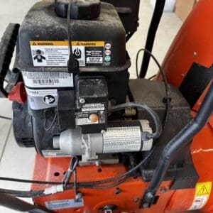 Gas-powered engine mounted on an orange tracked machine, with fuel cap, wiring, and warning labels visible