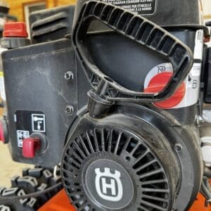 Close-up of a small gasoline engine with a ribbed pull-start housing and a red control knob on an orange machine; label text visible on the engine cover.