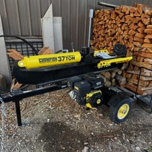 Yellow Champion 37-ton log splitter on a trailer beside a stack of cut firewood, outdoors by a metal building.