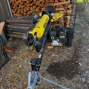 Gas-powered log splitter on a trailer sits in front of a stacked pile of firewood, ready for use or transport, with a tow hitch and safety chain in the foreground.
