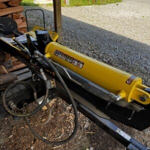 Yellow hydraulic log splitter mounted on a trailer with hoses, wood pile on the left, and a gravel driveway in a rural outdoor setting.