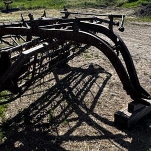 Rusty farming implement with curved metal arms and tines resting on dirt, casting shadows on the ground.