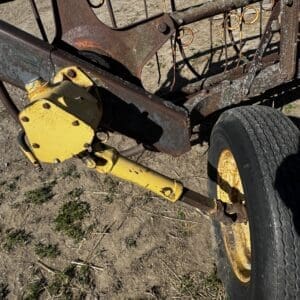 Rusty farm implement frame with a yellow hitch and wheel on dirt ground, showing worn metal and rusted connections.