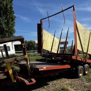 Red flatbed trailer with tall, yellow plywood panels standing upright on a rural lot, rusty metal frames and a two-axle wheel setup visible in the foreground.