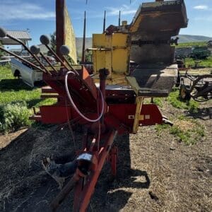 Red and yellow farm implement on dirt with a cat nearby, under a blue sky at a rural yard edge.