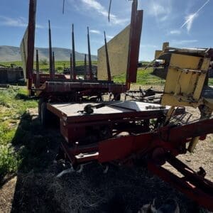 Abandoned red farm equipment with a yellow hopper in a sunlit field and distant hills; a small dog nearby.
