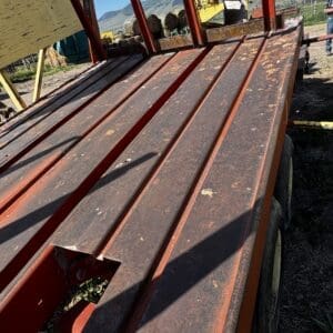 Rusted red metal farm trailer with long beams, outdoors on dirt with mountains in the background; partial shadow of a person on the right.