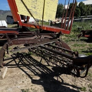 Rusty old hay rake with curved iron frame and tines, resting on wooden blocks in a rural yard.