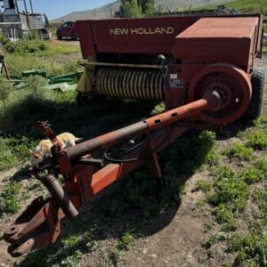 Red New Holland farm baler parked on a dirt lot with a curious cat nearby at the base of the hitch.