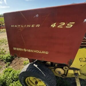 Red Hayliner 425 bale wagon with a yellow frame and wheel, on a grassy field under a clear blue sky.