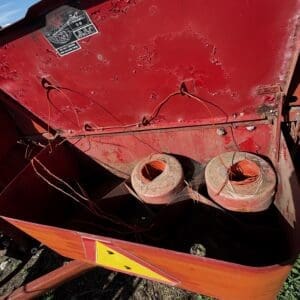 Red metal trailer with exposed orange wires and two spool-like cable reels, outdoors on dirt, warning triangle on the side