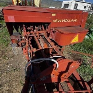 Red New Holland farm implement in a field, showing exposed linkage and hydraulic hoses under a clear blue sky with mountains in the background.