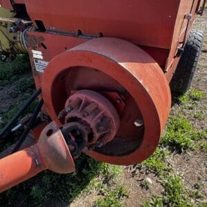 Close-up of a red farm implement showing the PTO drive shaft and circular housing attached to a trailer on dirt and grass.