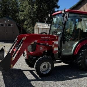 Red Mahindra compact tractor with front-end loader and enclosed cab on a gravel surface near a shed and greenhouse area, sunny day