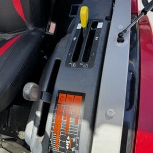 Close-up of a tractor's operator console showing two gear shift levers, a yellow handle, and an orange control label panel with symbols inside the cabin far from the seat edge.