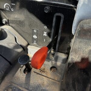 Close-up of dusty tractor cab floor showing a red safety lever and a black joystick-style control beside a gear selector panel.