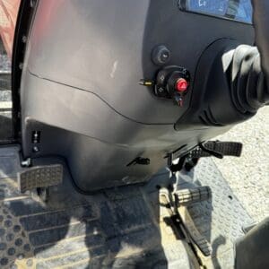Close-up of a heavy equipment operator’s cockpit showing pedals, floor mat, and dashboard with a red hazard switch.