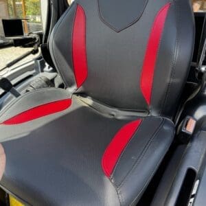 Driver seat in a vehicle with black perforated upholstery and red side panels; red logo on the headrest.