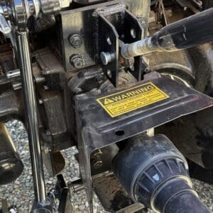 Close-up of a tractor rear hitch and PTO shaft area, showing mounting bolts, a metal bracket, and a yellow warning label about rotating parts.