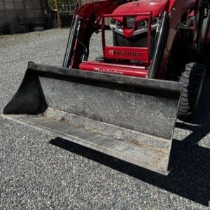 Red Mahindra compact tractor with a front-end loader bucket parked on gravel outdoors, with a shed and trees in the background.