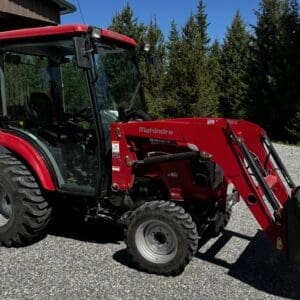 Red Mahindra tractor with a front loader and bucket, parked on gravel near trees.