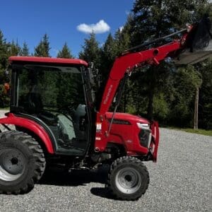 Red tractor with a front-end loader parked on gravel, forested background under a blue sky.