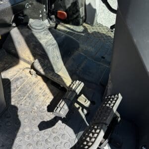 Foot pedals inside a vehicle cab (likely heavy machinery): two large pedals on a textured rubber floor mat, with a dusty surface nearby and an orange lever in the background.