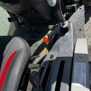 Inside a heavy equipment cab: joystick control with a red button, a black seat with a red stripe, and a 'To Start Engine' label on the console.