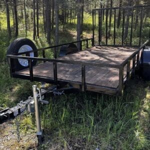Black utility trailer with a wooden flatbed and metal railing parked in a forest, showing the hitch, chains, and spare tire mounted on the side.