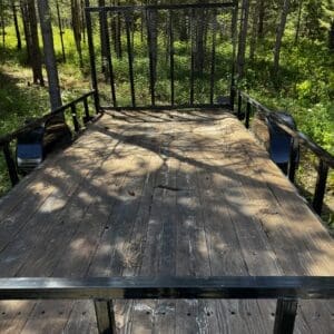 Wooden flatbed trailer in a forest, with a metal gate at the back and surrounding trees and greenery.