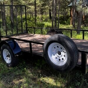 Black utility trailer with a wooden deck parked in a grassy, forested area.