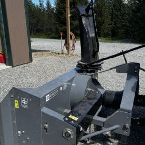 Heavy gray IronCraft skid-steer attachment on gravel, featuring a hydraulic arm and rotating chute, parked beside a shed with trees in the background.
