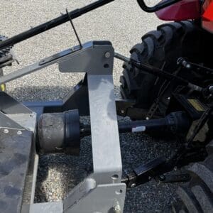 Rear three-point hitch on a tractor with hydraulic lift and PTO shaft, viewed from the side on a gravel surface, with a large tire in the background.