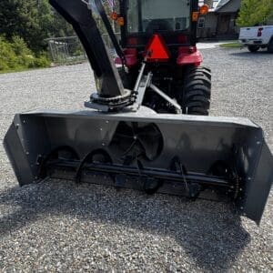 Red Mahindra tractor with a large front-mounted snow blower/auger attachment on a gravel driveway, mountains and trees in the background.