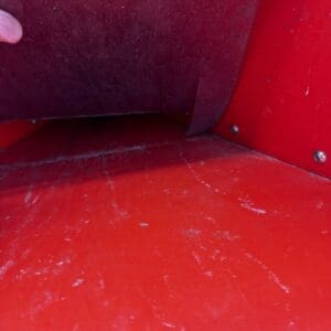 Close-up of a red metal surface with a brown flap being lifted by a finger; visible scratches and a small warning sticker in the corner.