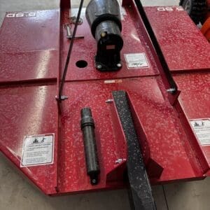 Close-up of a red automotive lift base in a workshop, featuring a black drive shaft and hitch-like component in the center with warning labels visible.