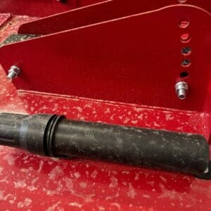 Close-up of red metal machinery with multiple drilled holes, bolts, and a black cylindrical rod resting on the surface.